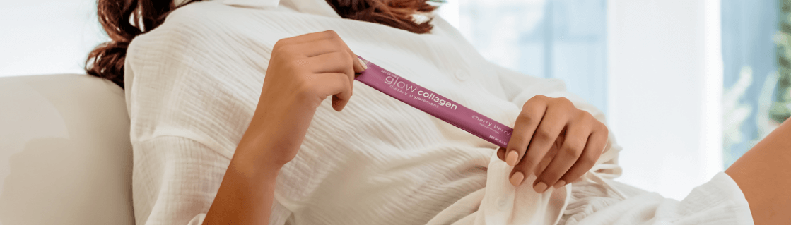 A young woman wearing white leaning back on a couch while holding a stick pack of AdvoCare Glow Collagen