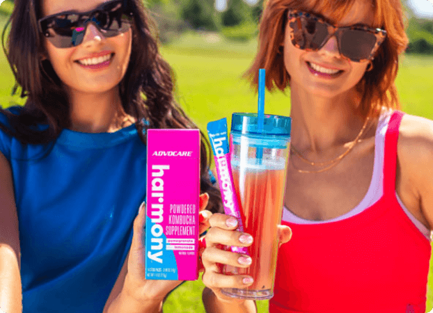 Two women standing side by side in a park, one holding the Harmony box and the other one holding a cup with pre mixed Harmony and a stick pack.