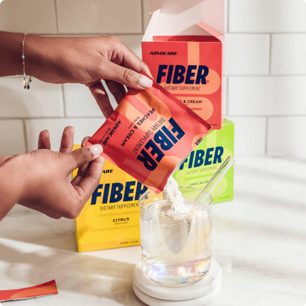 Hands pouring a packet of Peach Fiber into a glass of water on a kitchen counter in front of more boxes of AdvoCare Fiber.